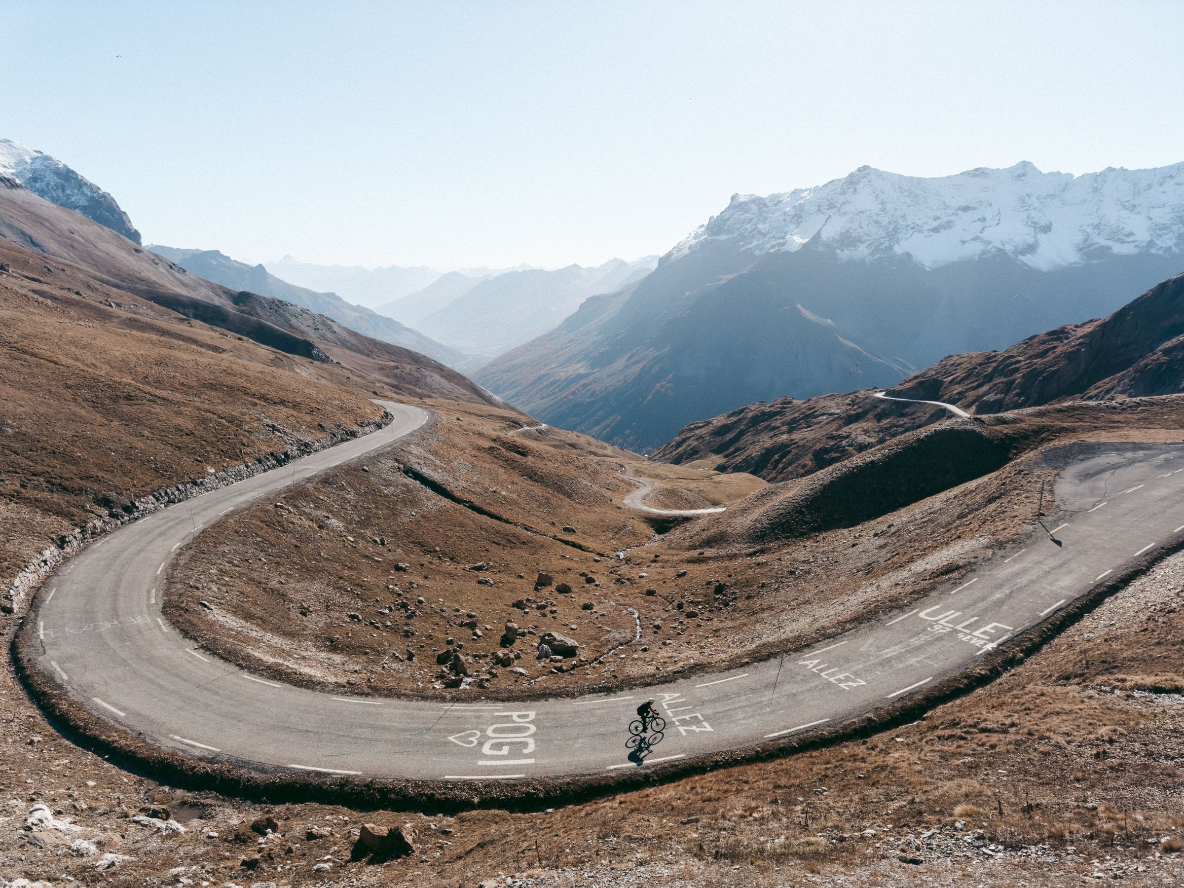 A lone cyclist rounds a hairpin on a high alpine pass. Fans have painted ALLEZ and POGI on the tarmac, and snow-covered peaks rise over a hazy valley beyond.